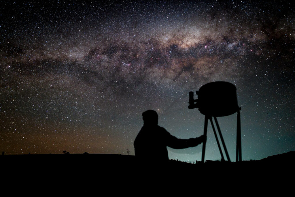 The silhouette of a person beside a large dobsonian telescope on a dark night with a sky above showing many stars and the Milky Way above