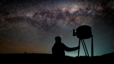 The silhouette of a person beside a large dobsonian telescope on a dark night with a sky above showing many stars and the Milky Way above