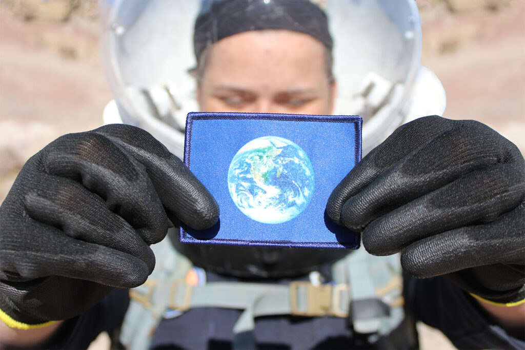 A flag of the Earth being held by an analog astronaut in the desert of Utah. The astronaut is wearing a spacesuit with a clear helmet visor and has black gloves. The image highlights the symbol of Earth on a blue background.