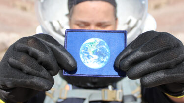 A flag of the Earth being held by an analog astronaut in the desert of Utah. The astronaut is wearing a spacesuit with a clear helmet visor and has black gloves. The image highlights the symbol of Earth on a blue background.